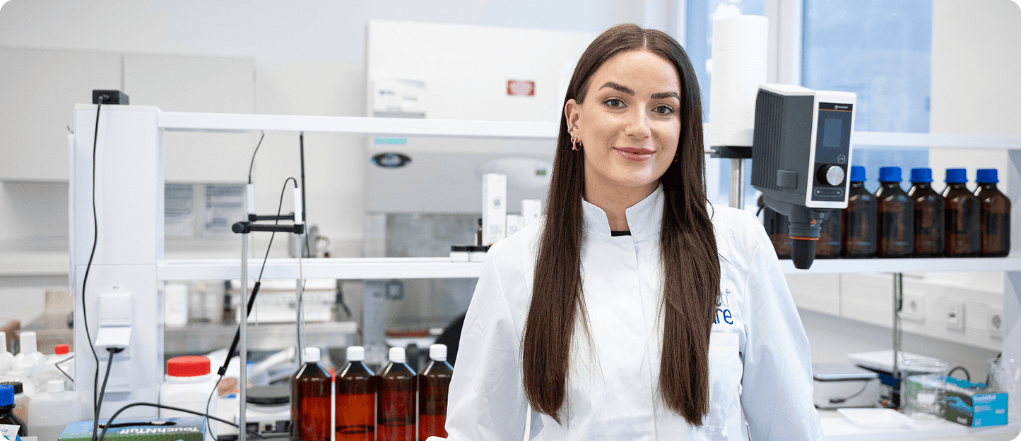 Eine Frau in einem weißen Laborkittel steht lächelnd in einem modernen Labor, umgeben von wissenschaftlichen Geräten, braunen Glasflaschen und Laborbedarf.