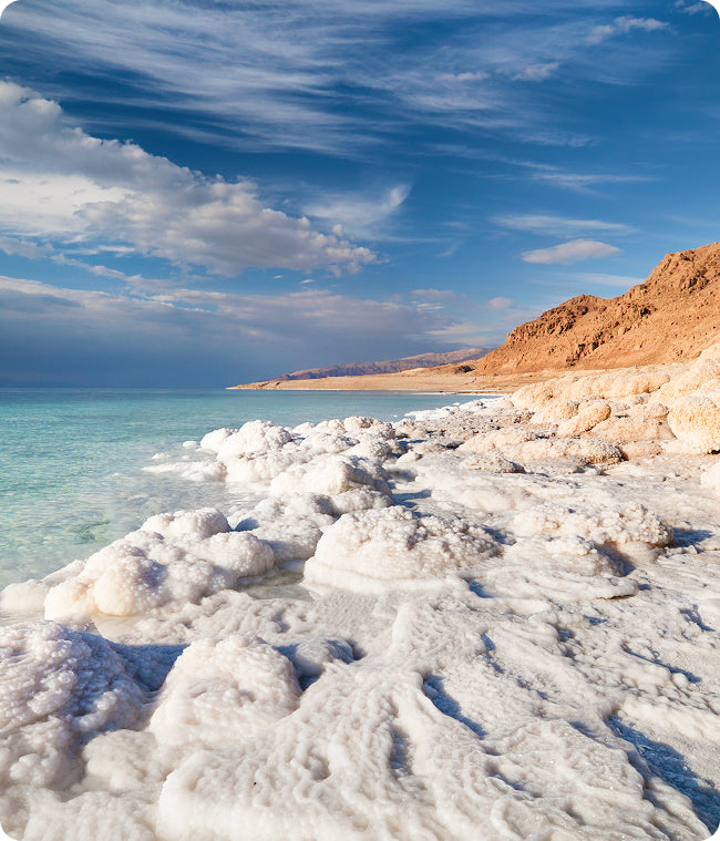 Eine felsige, mit weißen Salzformationen bedeckte Küste mit türkisfarbenem Wasser unter einem blauen Himmel mit vereinzelten Wolken; im Hintergrund erheben sich rötlich-braune Hügel entlang der Küste.
